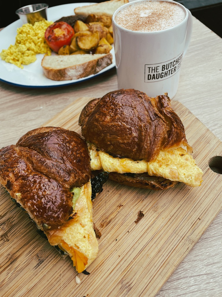 A croissant and egg sandwich cut in two halves and placed on a pale wooden cutting board. The croissants are glistening with butter. The egg is dripping with cheese. To the right of the cutting board is a white mug filled with chai. On the mug is the name of the restaurant "The Butcher's Daughter". In the background is a platter of vegan "eggs" potatoes, fried tomato, and hearty country bread.