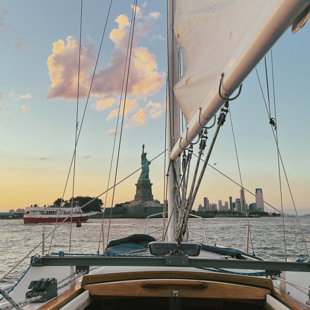 The Statue of Liberty stands defiantly in the background as the deck and sail of a small boat frames her. On the bottom left is a small red ferry and on the right is New Jersey's skyline. This was taken at sunset.
