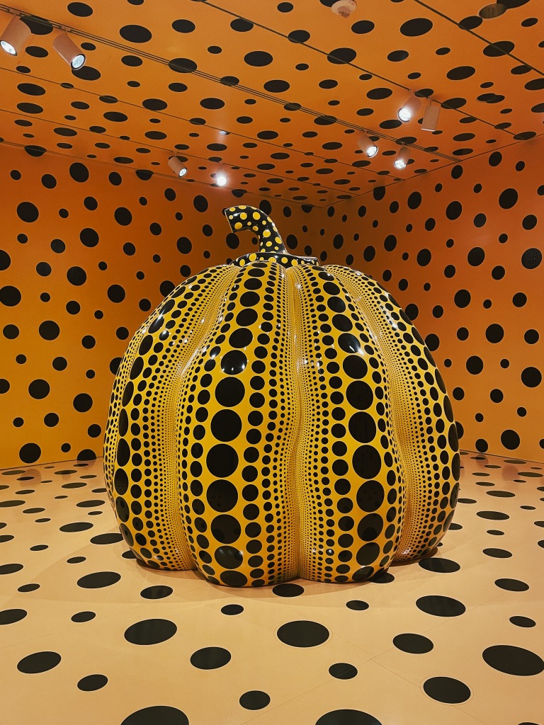 Yayoi Kusama at the Hirschorn museum in Washington D.C. In a bright orange room with black polka dots painted on the walls, ceiling, and floor, there is a yellow pumpkin. The pumpkin is sunken into the floor so the bottom is cut off. Painted on the pumpkin are black polka dots in various sizes.