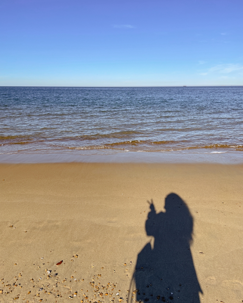 The Atlantic ocean is receding. The sky is a gradient of deep blue, light blue, and hazy gray to mark the horizon. The bottom half of the photo is densely packed and wet sand. There is a shadow of a person holding up a peace sign.