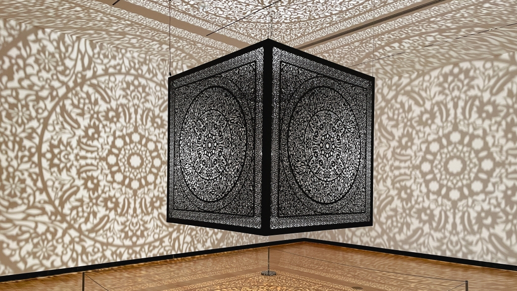 All Flowers Are for Me (2019) by Anila Quayyum Agha (b. 1965). A black metal cube with an intricate kaleidoscope pattern cut-out on all sides. In the cube is a light that creates shadows on every surface.