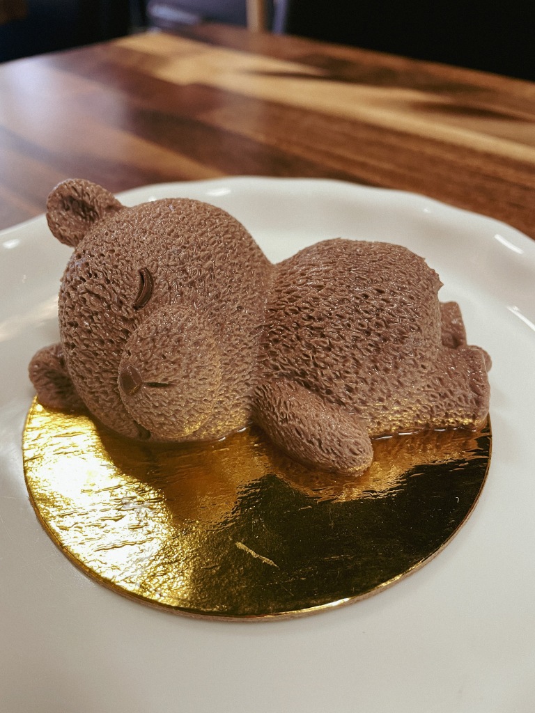An incredibly textured chocolate mousse dessert in the shape of a cartoon bear laying on its stomach. It is laying on a gold round disc on top of a white ceramic dish.